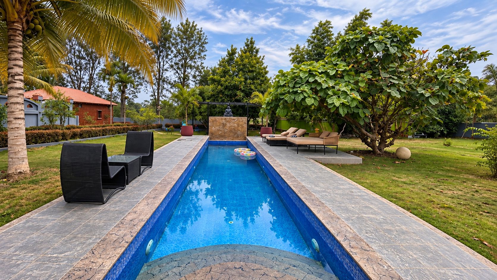 A rectangular swimming pool with a stone border, surrounded by a patio with outdoor furniture, and lush greenery in the background.