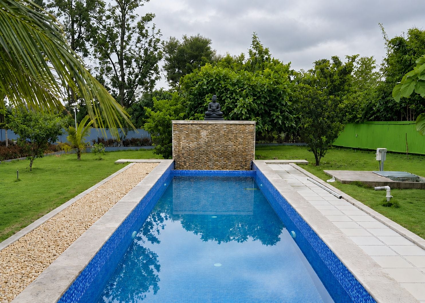 Piscina retangular com muro de pedra e uma estátua no centro, cercada por um gramado bem cuidado e árvores.
