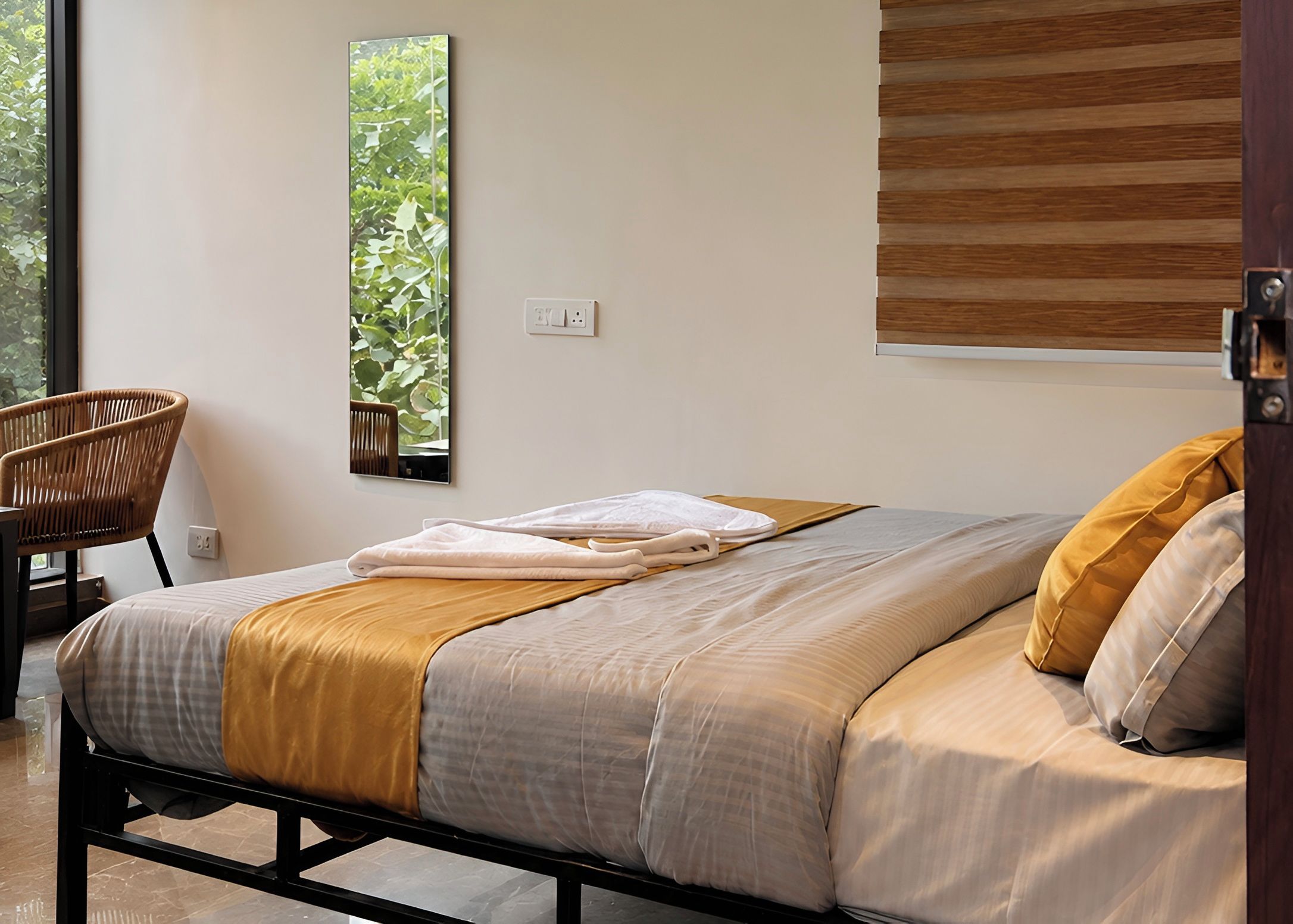 A neatly made bed with a grey and yellow bedspread, a wooden chair, and a large window with a view of greenery.
