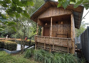 A bamboo structure with a balcony, surrounded by greenery and a small water feature.