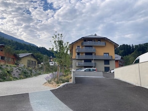 A multi-story building with balconies, a parking area, and a mountainous backdrop. - Les Pics Alpins (Saint Gervais)