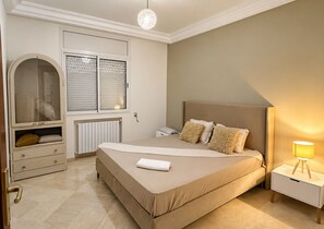 A bedroom with a bed, bedside table, lamp, wardrobe, and a window with blinds. - The Quiet Corner (Tunis)