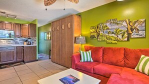 A compact living space with a kitchenette, a red sofa, and tropical-themed wall art. - AVAILABLE IRONMAN 2026 - OCEANVIEW Brilliant condo Close to the PIER For Ironman (Kailua-Kona)