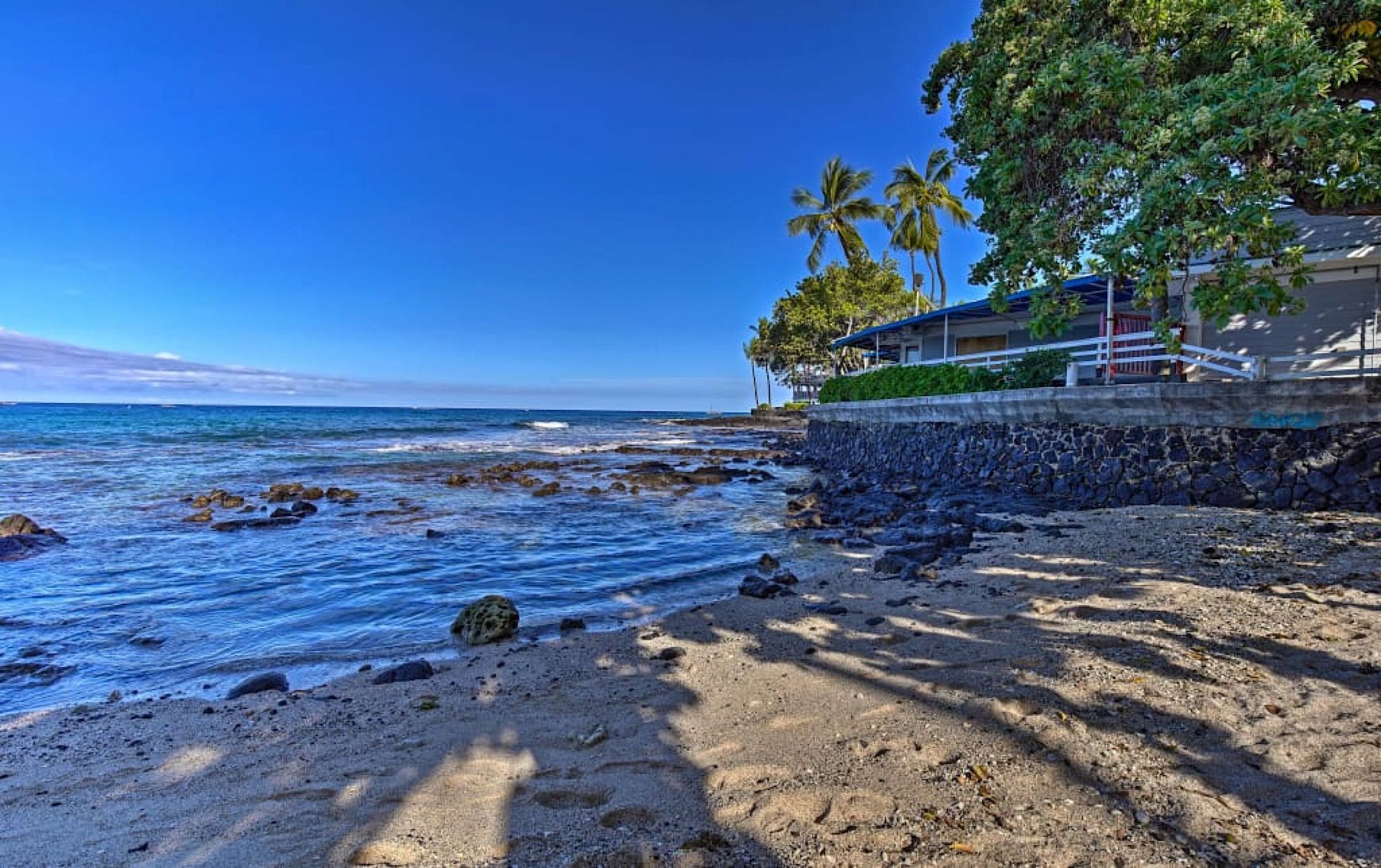 A beach with a stone wall, palm trees, and a building with a blue roof.