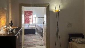 Ein Zimmer mit Bett, Nachttisch, Lampe und Blick in einen anderen Raum mit Fenster und Vorhängen.