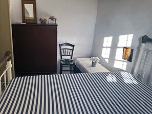 A room with a striped bed, a wooden wardrobe, a chair, a small table with a vase, and a window with a picture.