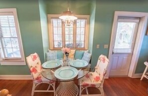 Comedor con mesa de vidrio rodeada de sillas con estampado floral, candelabro y ventana con persianas.