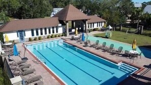 Una piscina rectangular con tumbonas y sombrillas, rodeada por un pabellón cubierto y un edificio con techo de tejas.