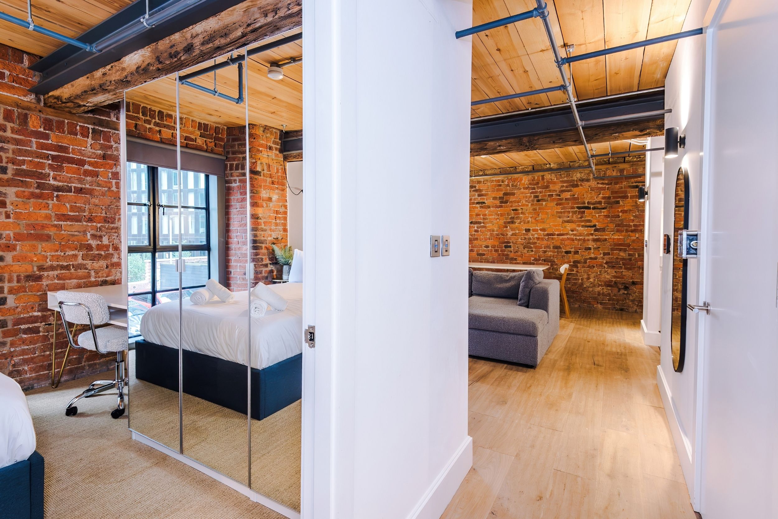 A modern room with exposed brick walls, a large window, a bed with a white headboard, and a small seating area with a gray sofa.