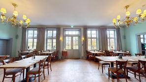 En rummelig spiseafdeling med flere borde og stole, store vinduer med gardiner og lysekroner.
