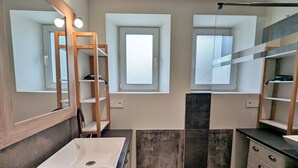 Une salle de bain moderne avec un grand miroir, deux fenêtres, un lavabo sur colonne et des étagères intégrées.