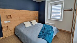 Une chambre avec une tête de lit en bois, un lit avec une literie grise, un plaid bleu, une table de chevet et une fenêtre avec des stores.