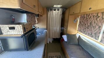 A compact RV interior with a kitchenette, a seating area with cushions, and a window with curtains.