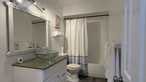 Une salle de bain avec un lavabo en verre, un meuble-lavabo blanc, des toilettes, une baignoire et un rideau de douche.