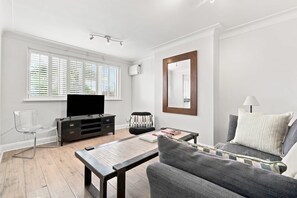 A modern living room with a flat-screen TV, a sofa, a coffee table, and a large window with blinds. - Modern Apartment with WiFi & Kitchen Near Train Station (Hutton)