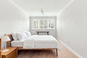 Ein Schlafzimmer mit einem Bett, einem Nachttisch mit Lampe, einem Fenster mit Jalousien und einer Deckenleuchte.