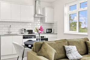 A modern kitchen with white cabinets, a stainless steel range hood, and a dining area with a green sofa and a vase of flowers.