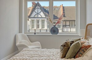 Een slaapkamer met een bed, een stoel, een raam met uitzicht op een gebouw en een decoratieve ventilator.