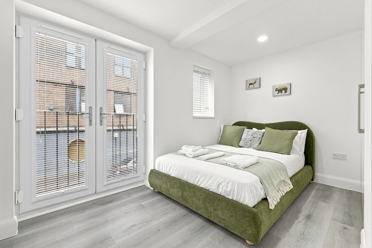 Une chambre à coucher avec un lit recouvert d’un tissu vert, du linge de lit blanc et une grande fenêtre avec des stores.