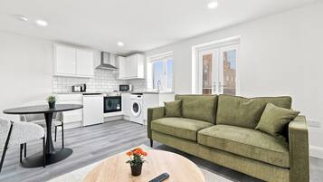 Eine moderne Küche mit weißen Schränken, ein Essbereich mit einem runden Tisch und ein grünes Sofa in einem Wohnzimmer.