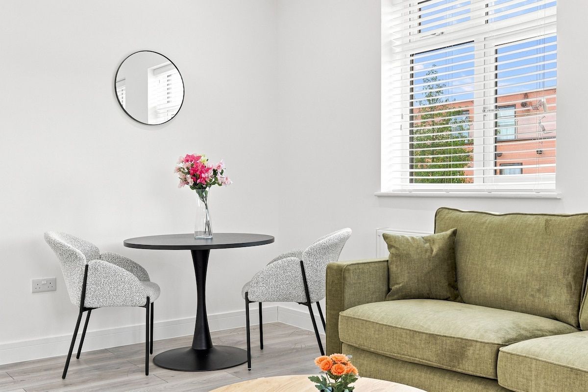 A modern living room with a round table, two chairs, a green sofa, and a window with blinds.