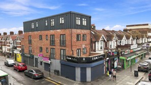 A street view with a row of terraced houses, a modern building with a sign for 'SN Jewellery', and parked cars. - Brand New 1-Bed,  Ilford Lane, Near Station (Greater London)