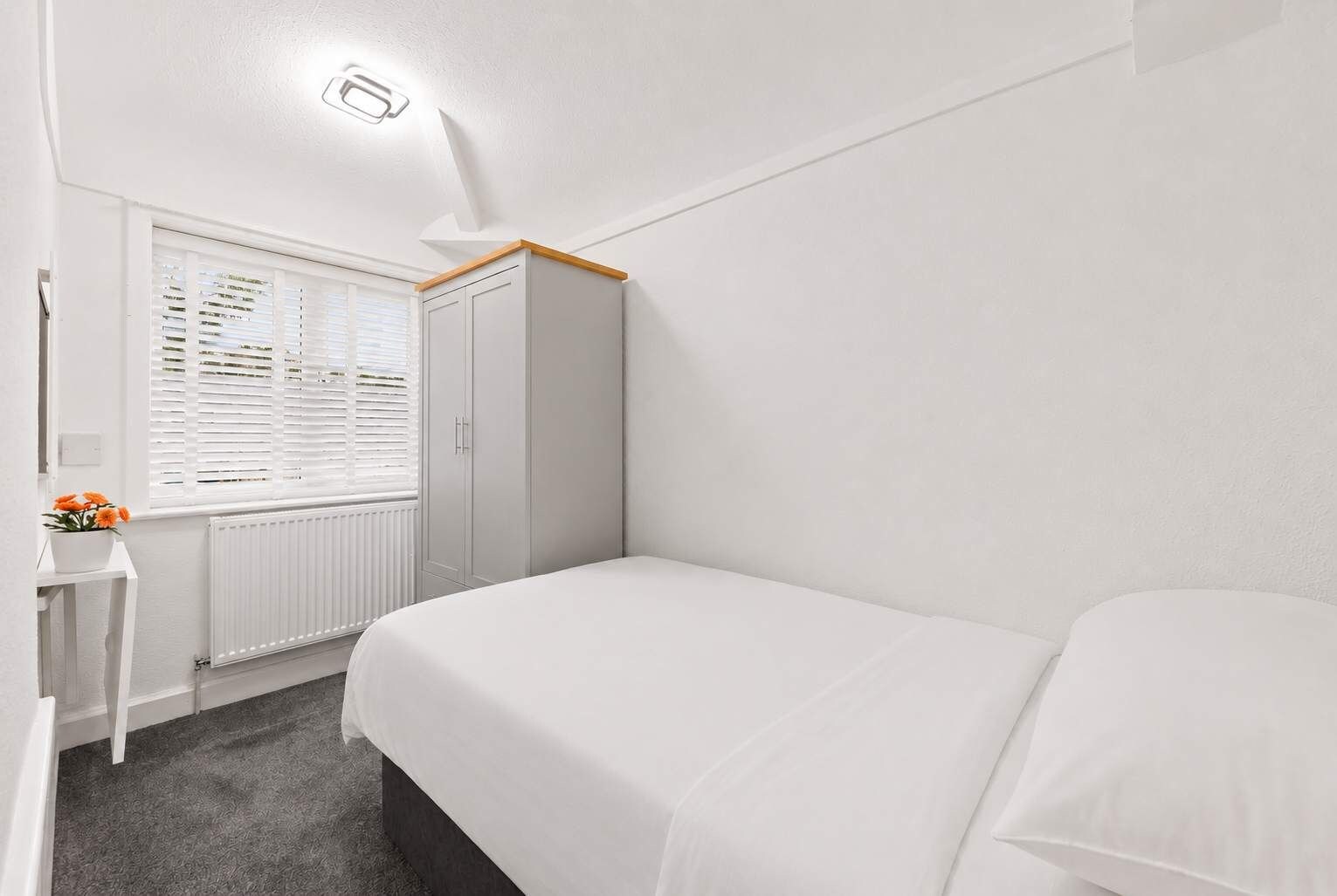 A bedroom with a bed, a wardrobe, a radiator, a window with blinds, and a small table with flowers.