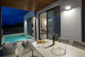 Un espacio moderno al aire libre con zona de comedor, piscina, mesa con comida y vino, y vista hacia el interior.