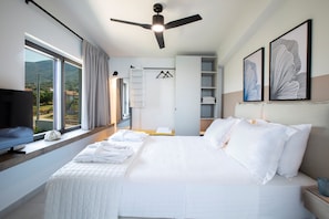 Habitación de hotel con cama, televisión, ventilador de techo, ventana con cortinas y una obra de arte en la pared.