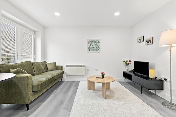 Een moderne zithoek met een groene bank, een ronde salontafel, een zwart tv-meubel en een vloerlamp.