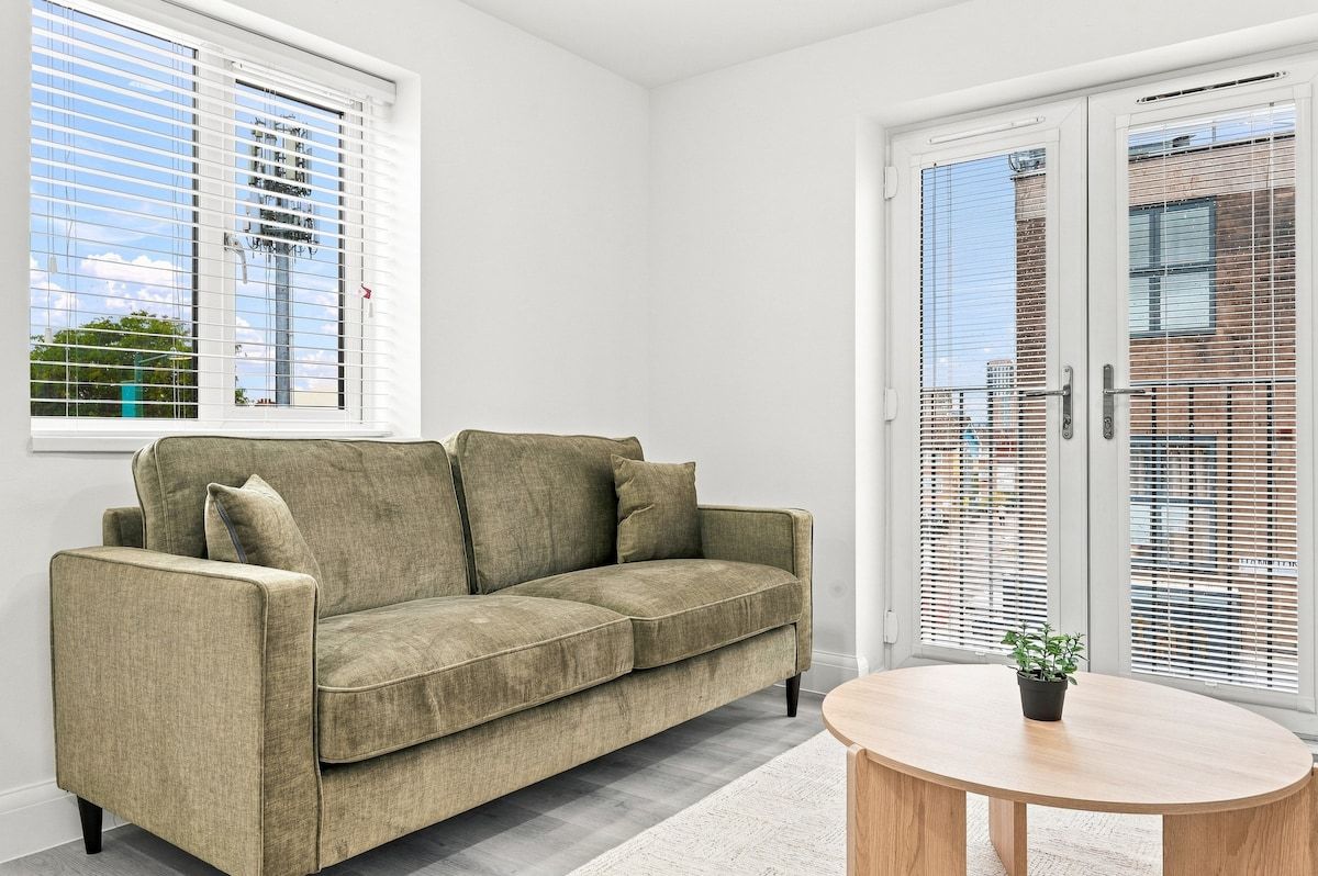 Ein Zimmer mit einem beigen Sofa, einem runden Holztisch und einem Fenster mit Jalousien.
