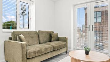 Ein Zimmer mit einem beigen Sofa, einem runden Holztisch und einem Fenster mit Jalousien.