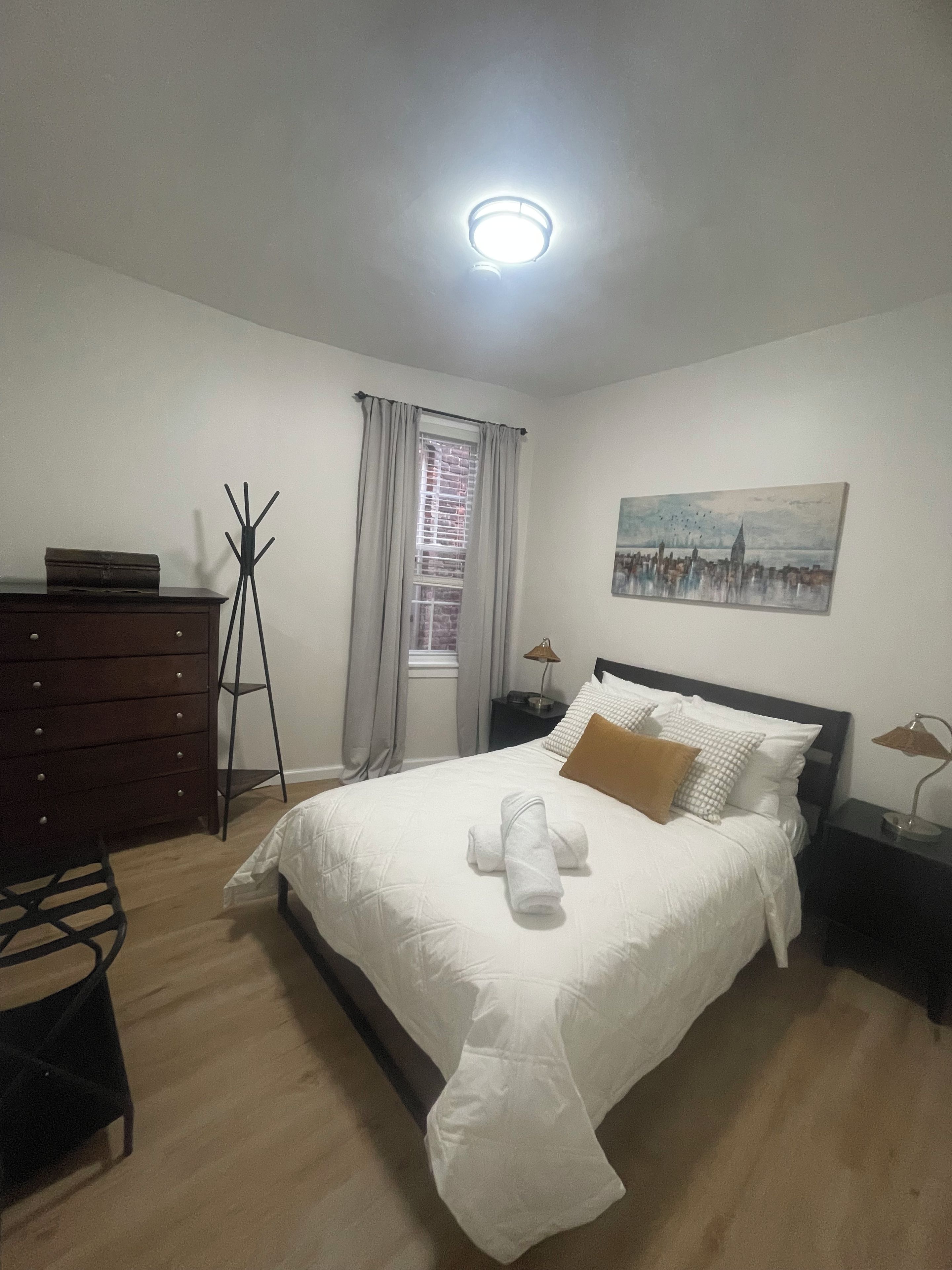 A bedroom with a bed, a dresser, a coat rack, a window with curtains, a nightstand, a lamp, and a painting on the wall.