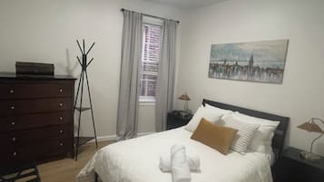 A bedroom with a bed, a dresser, a coat rack, a window with curtains, a nightstand, a lamp, and a painting on the wall.