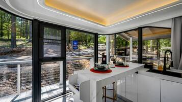 A modern kitchen with large windows, a white countertop, and a view of a forested area.