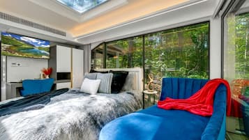 A modern bedroom with a large bed, a blue sofa, a skylight, and a view of greenery.