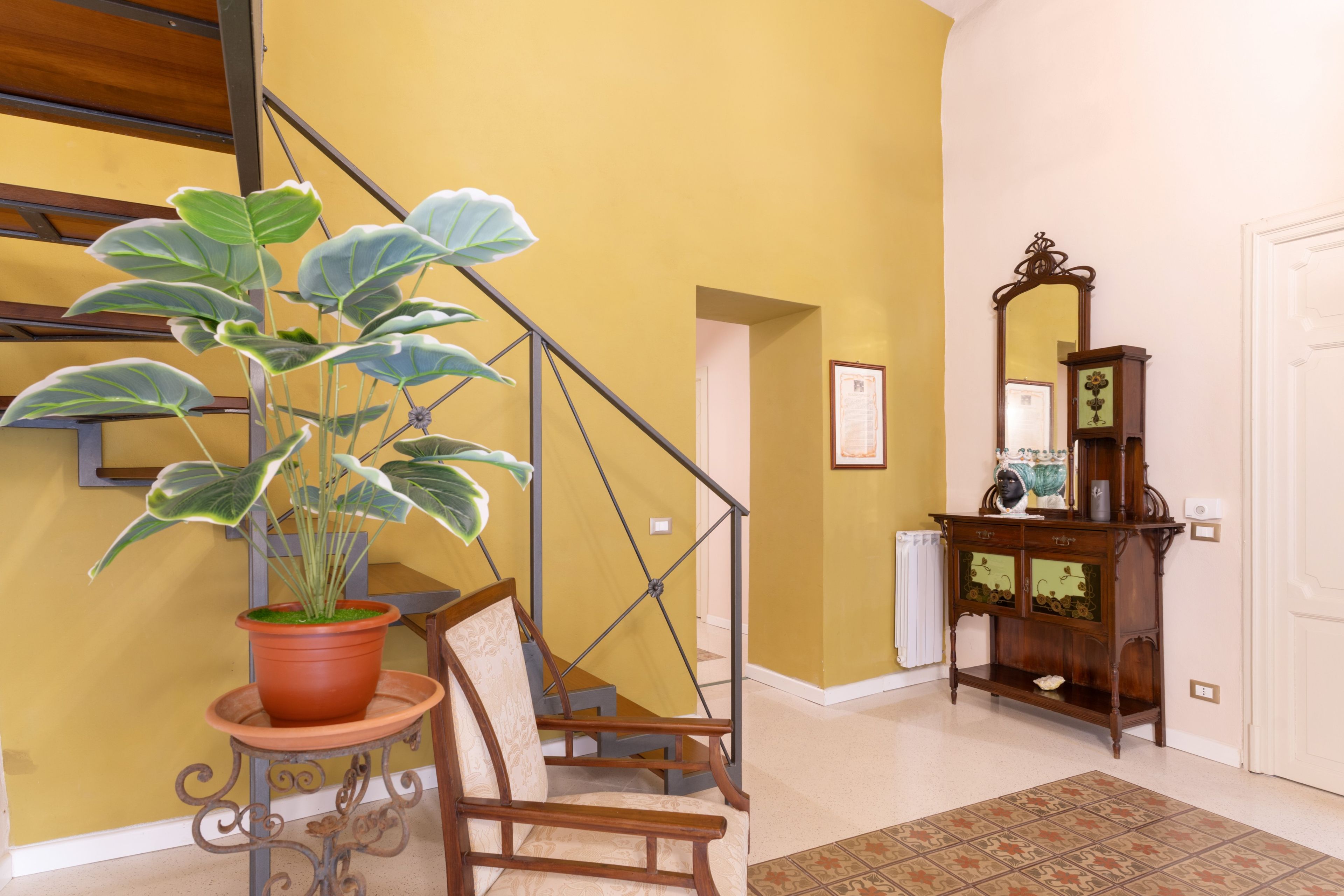 Una escalera con una planta en maceta, una silla, un mueble de madera con un espejo y una alfombra estampada.