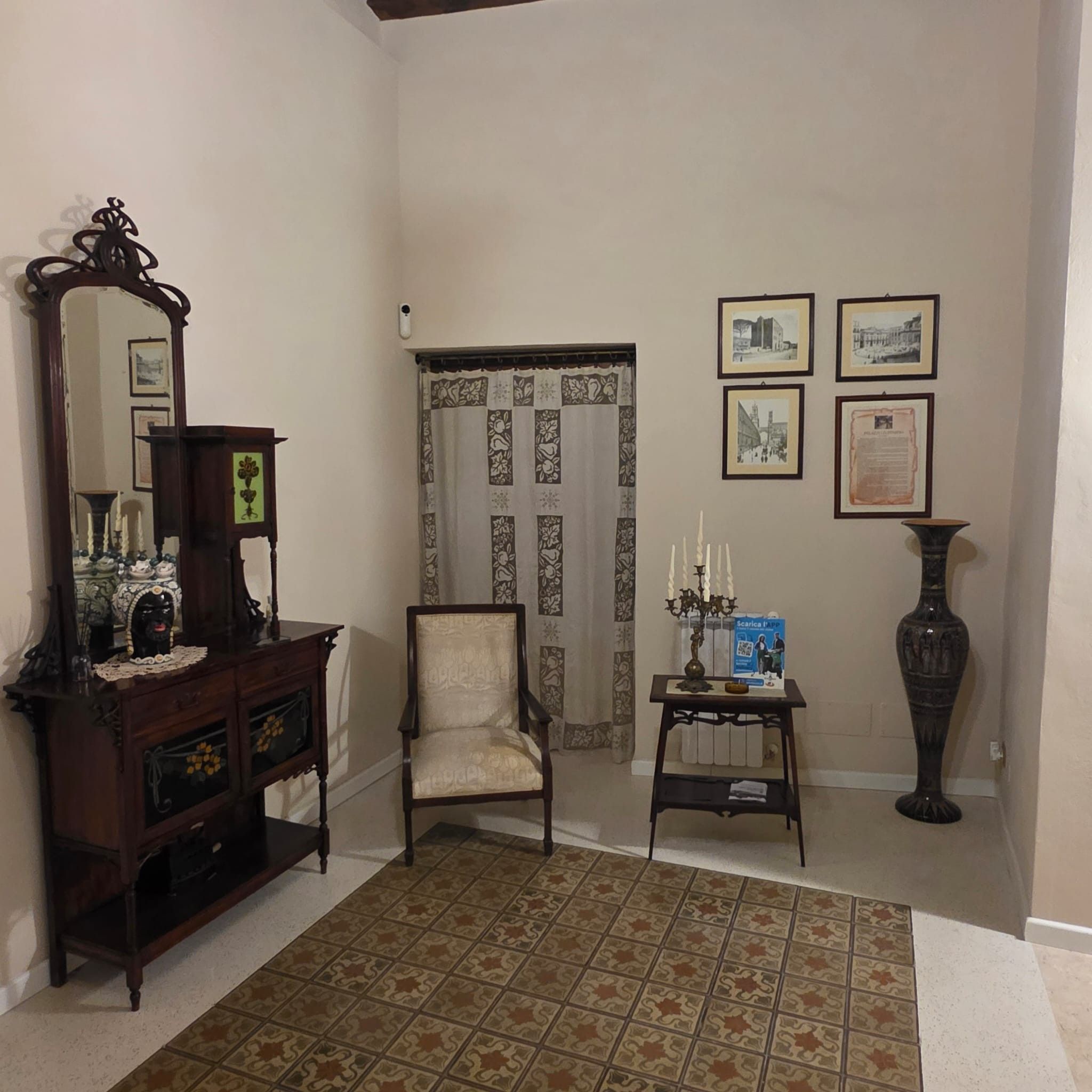 Una habitación con un aparador de madera, una silla, una mesita con un jarrón, un espejo y una puerta con cortinas estampadas.