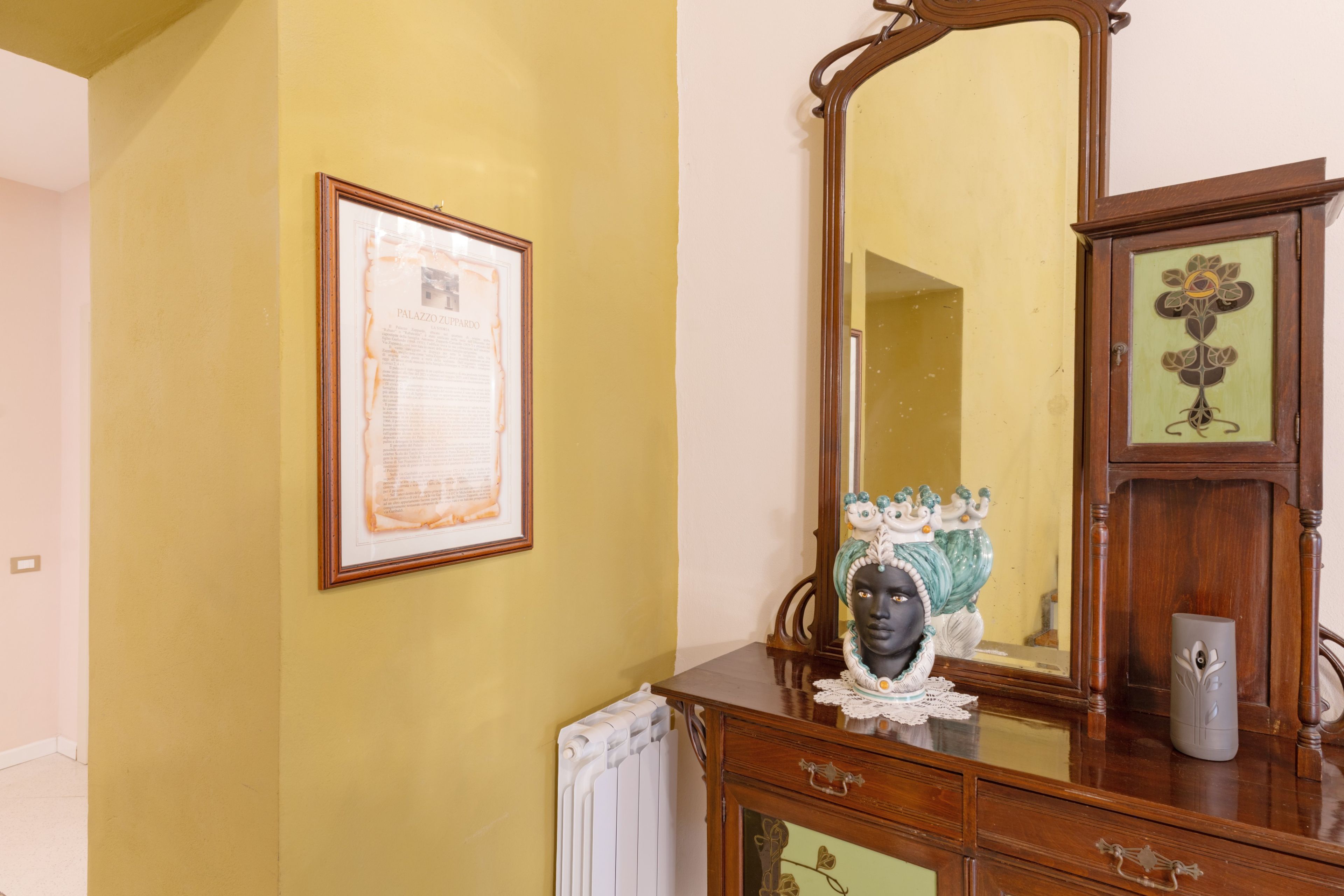 A room with a large mirror, a framed document on the wall, and a wooden cabinet with a decorative vase.
