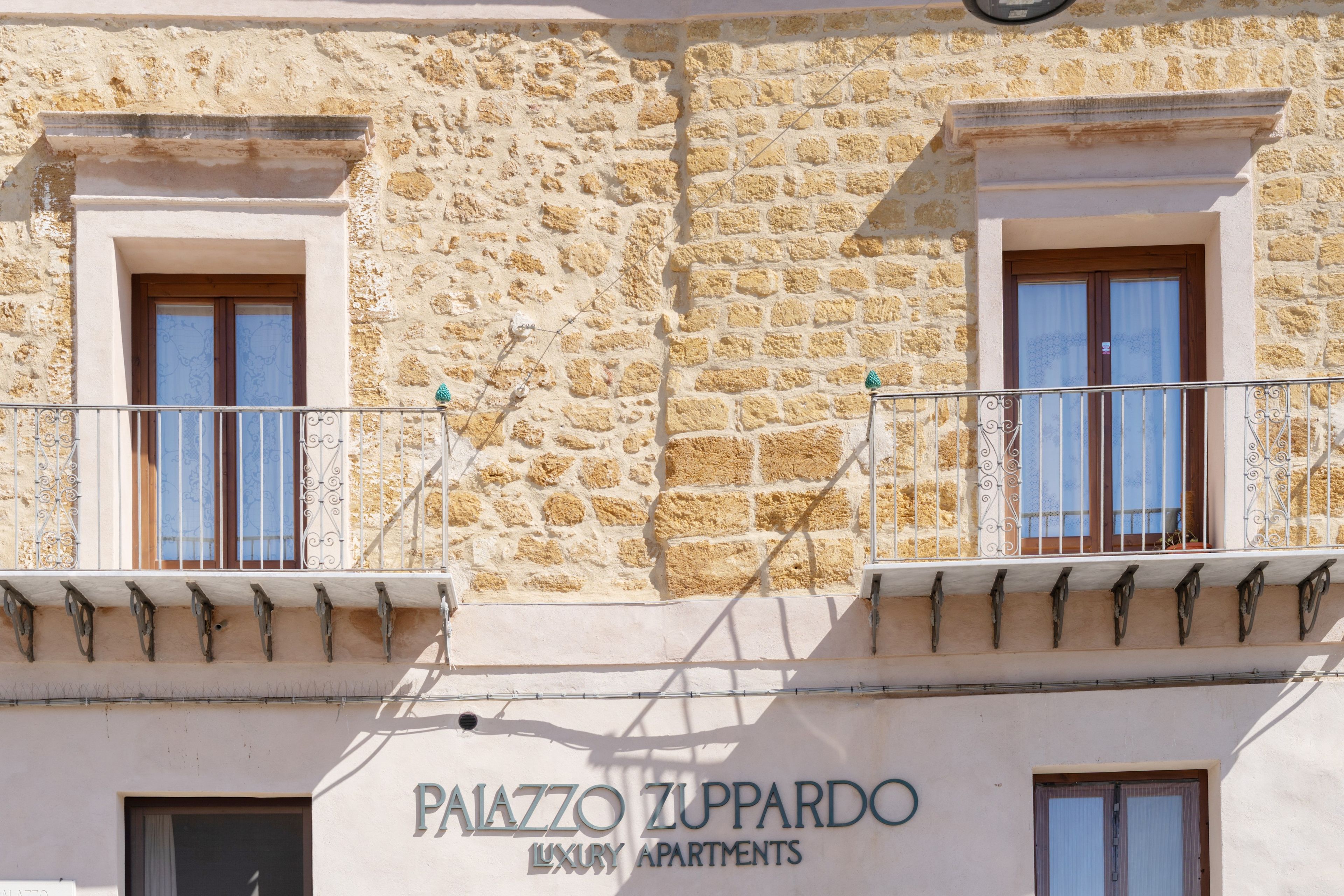 A building facade with 'PAIAZZO ZUPPARDO LUXURY APARTMENTS' signage, featuring stone walls, two balconies with metal railings, and windows with wooden frames.