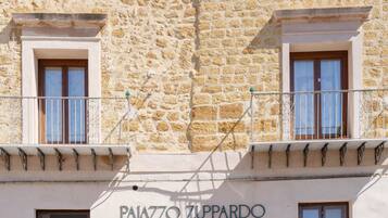 A building facade with 'PAIAZZO ZUPPARDO LUXURY APARTMENTS' signage, featuring stone walls, two balconies with metal railings, and windows with wooden frames.