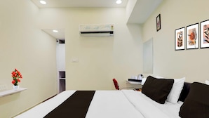 Habitación de hotel con cama, escritorio, silla, una silla roja, un jarrón blanco con flores rojas, aire acondicionado montado en la pared y cuadros en la pared.