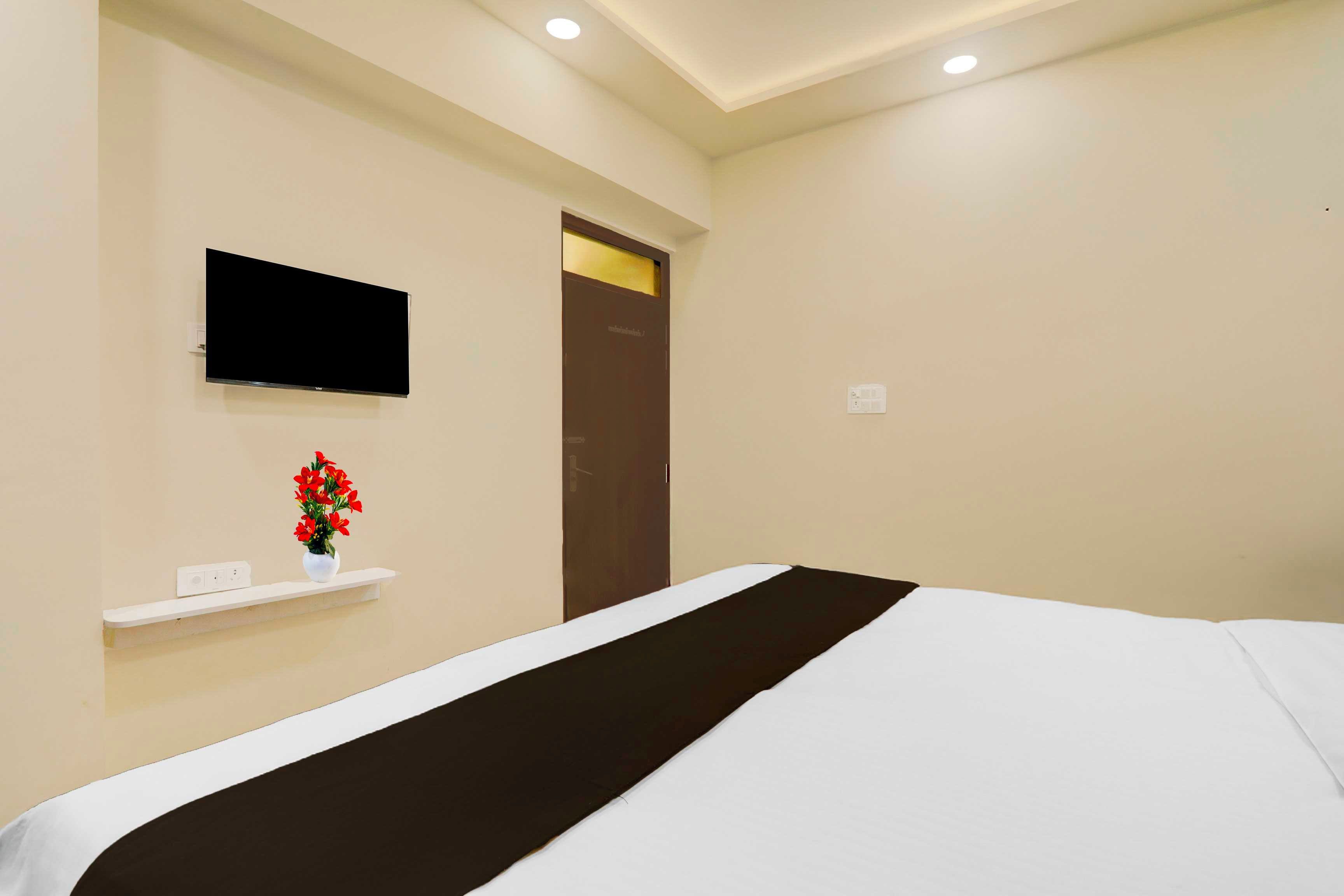 Una habitación de hotel con una cama, un televisor montado en la pared, un pequeño estante con un jarrón de flores y una puerta con pomo dorado.