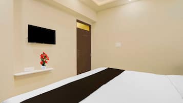 Una habitación de hotel con una cama, un televisor montado en la pared, un pequeño estante con un jarrón de flores y una puerta con pomo dorado.