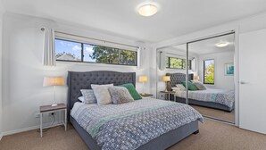 A bedroom with a large bed, two bedside tables, a mirror, and a window with a view of trees. - Beach Road Retreat  Pet Friendly Walk to Beach (Torquay)