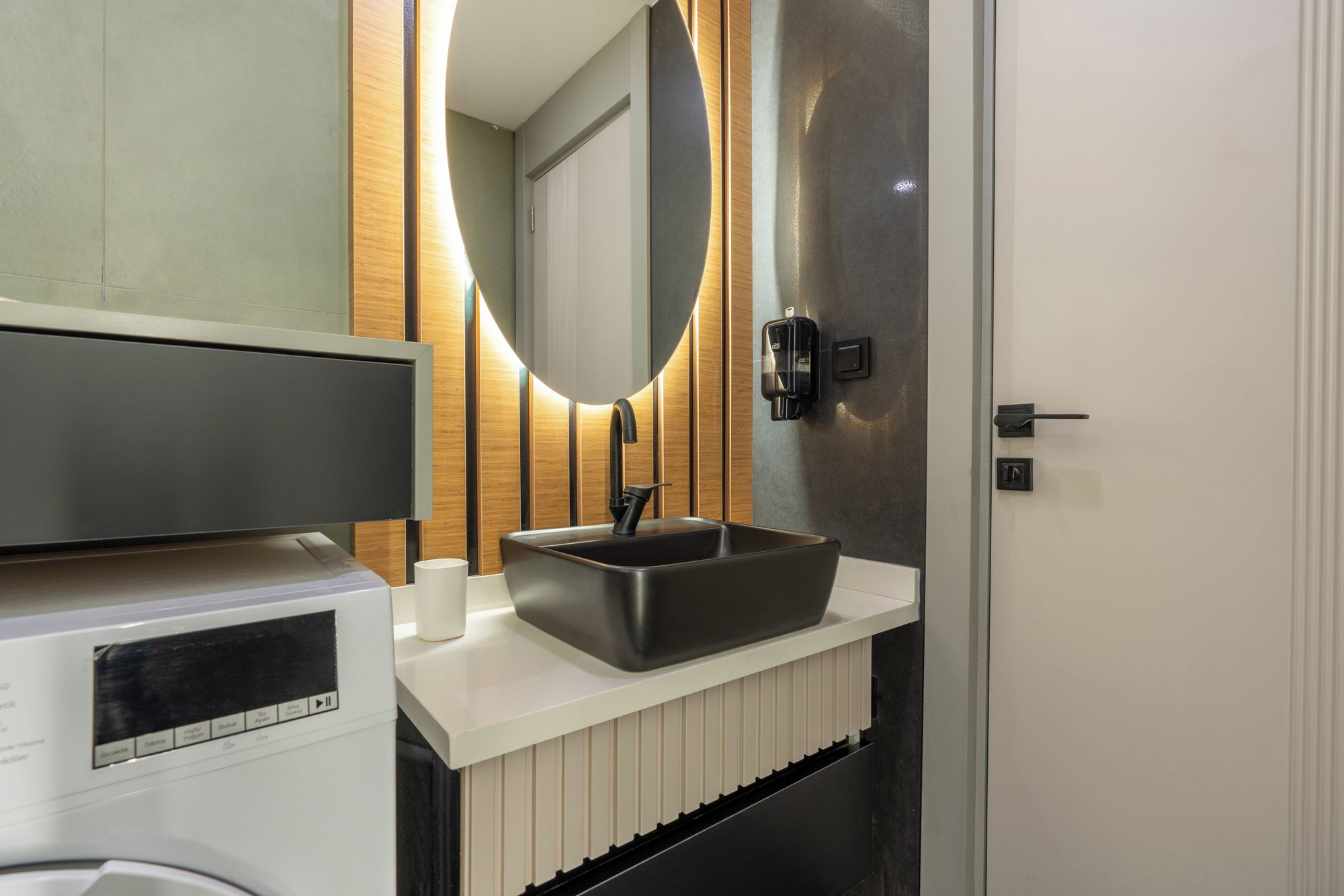 A modern bathroom with a built-in washing machine, a vessel sink, and a mirror with a wooden accent wall.