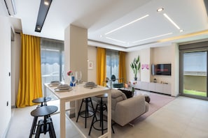 A modern living room with a sofa, a dining table with chairs, a TV mounted on the wall, and a large window with curtains.