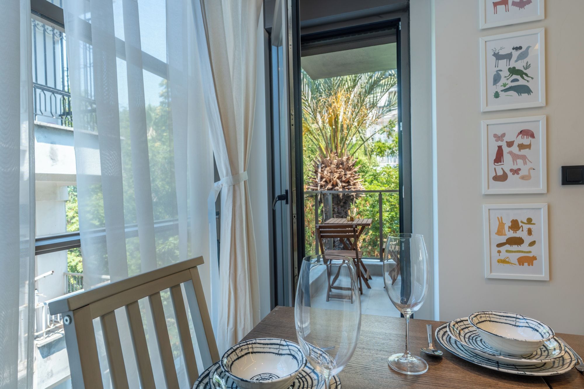 A dining area with a table set for two, a glass of wine, and a view of the outdoors through a window.
