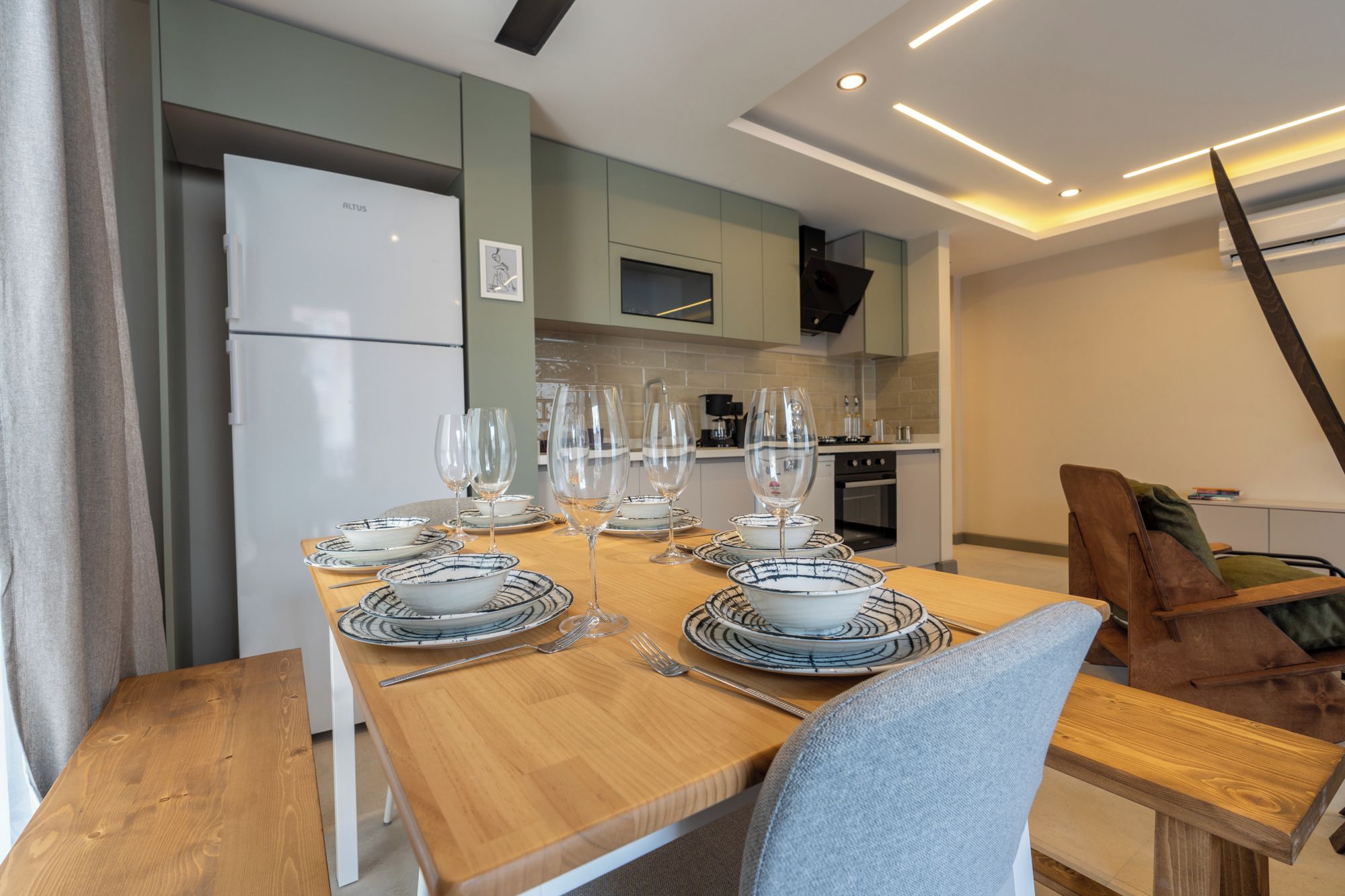 A modern kitchen with a dining area set for six people, featuring a wooden table and chairs, a white refrigerator, and a kitchen counter with appliances.