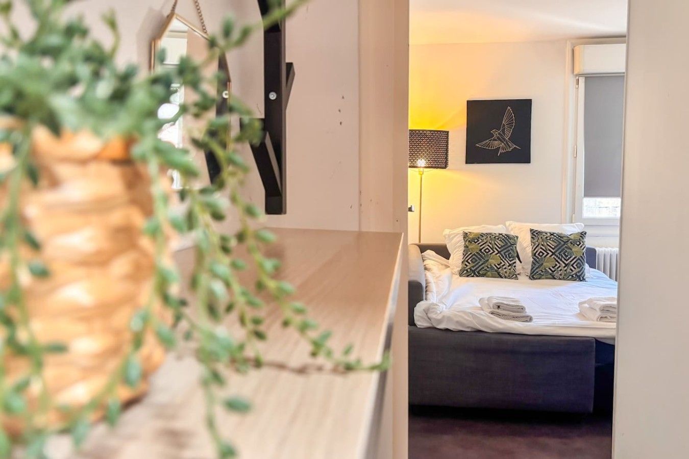 Quarto de hotel com cama, almofadas, uma obra de arte na parede e uma planta.